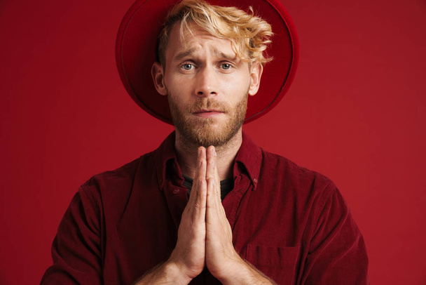 White bearded man wearing hat frowning while making prayer gesture isolated over red wall - Foto, Imagem
