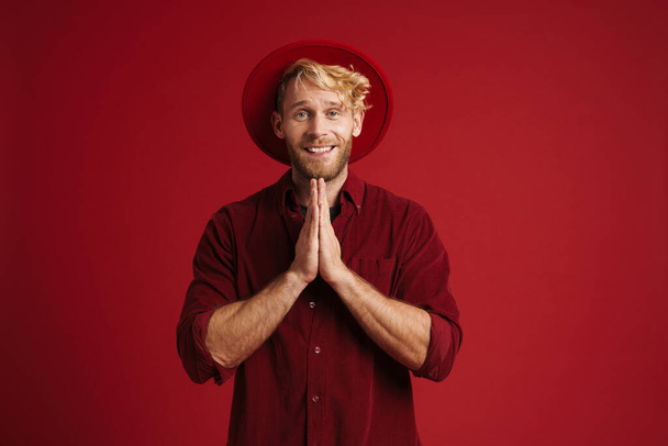 White bearded man wearing hat smiling and holding hands together isolated over red wall - Foto, immagini