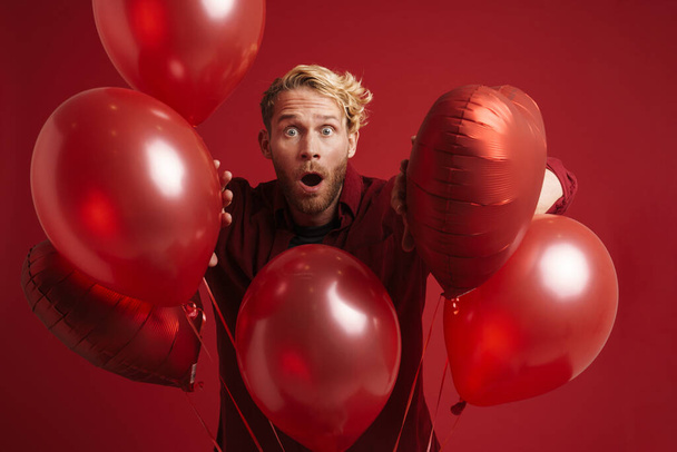 Bearded blonde man expressing surprise while posing with balloons isolated over red background - Foto, imagen