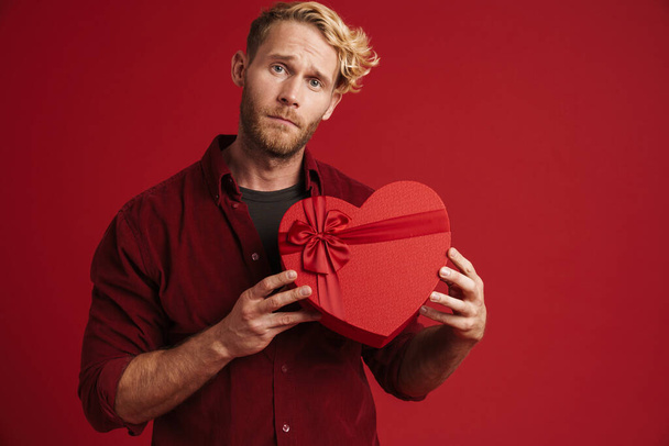 brodaty blondynka mężczyzna marszczenie brwi pokazując serce prezent pudełko izolowane na czerwonym tle - Zdjęcie, obraz