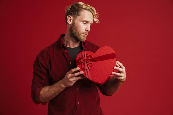 Bearded blonde man frowning while showing heart gift box isolated over red background - Fotoğraf, Görsel