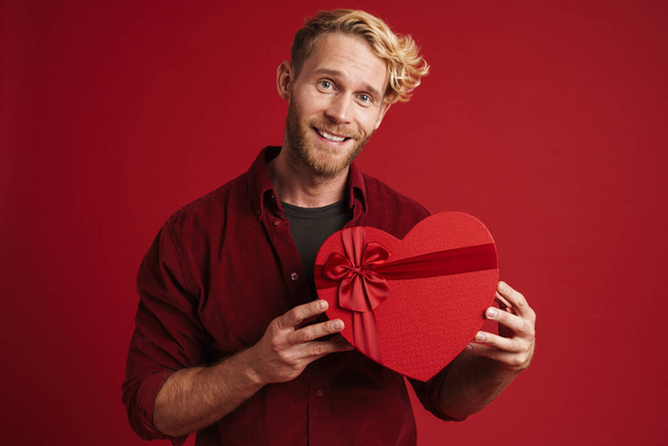 Bearded blonde man smiling while showing heart gift box isolated over red background - Φωτογραφία, εικόνα