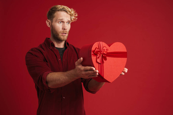 Bearded blonde man frowning while showing heart gift box isolated over red background - Photo, image