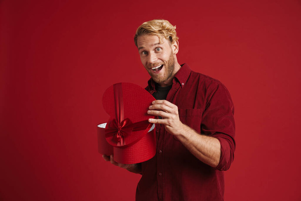 White man expressing surprise while opening heart gift box isolated over red wall - Фото, зображення