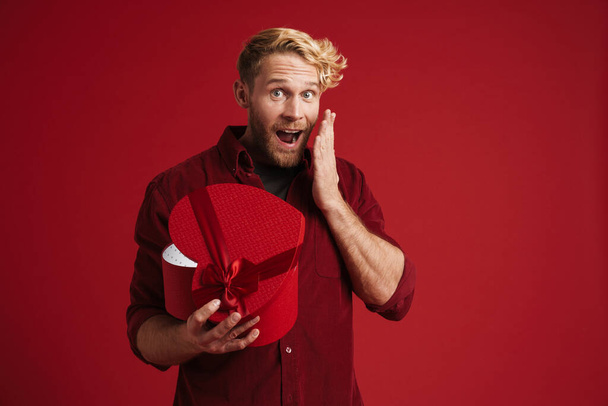White man expressing surprise while opening heart gift box isolated over red wall - Foto, immagini