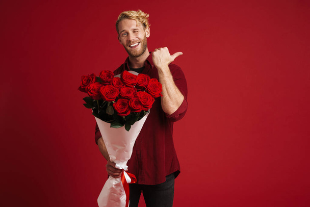 White bearded man pointing finger aside while posing with roses isolated over red wall - Фото, изображение