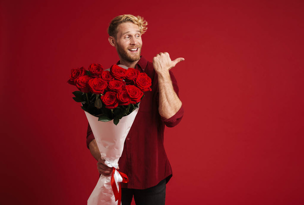 White bearded man pointing finger aside while posing with roses isolated over red wall - Photo, Image