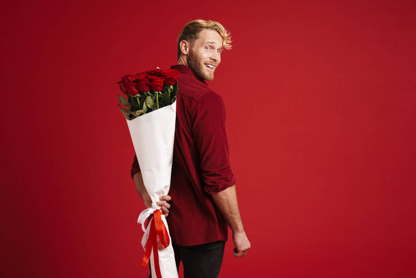 Blonde man smiling while posing with bouquet of roses isolated over red background - Foto, Imagem