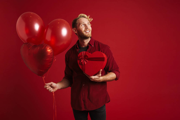 Bearded blonde man smiling while posing with balloons and gift box isolated over red background - Fotoğraf, Görsel