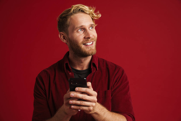 Bílý vousatý muž v košili s úsměvem a pomocí mobilu izolované přes červenou stěnu - Fotografie, Obrázek