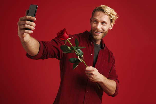 White bearded man holding rose while taking selfie on cellphone isolated over red wall - Foto, afbeelding