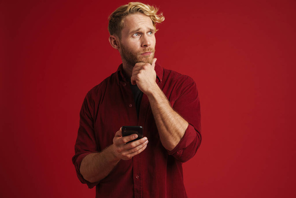 White puzzled man wearing shirt using mobile phone isolated over red wall - Foto, Bild