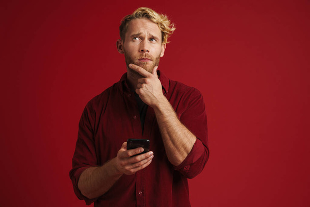 White puzzled man wearing shirt using mobile phone isolated over red wall - Φωτογραφία, εικόνα