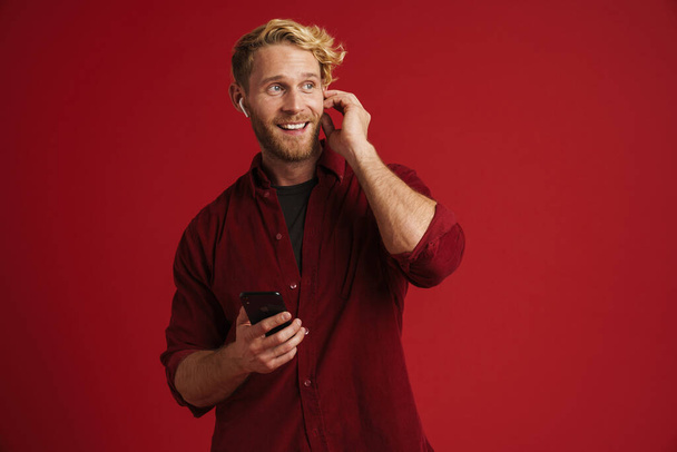 Blonde man listening music with earphones and using cellphone isolated over red wall - Fotó, kép