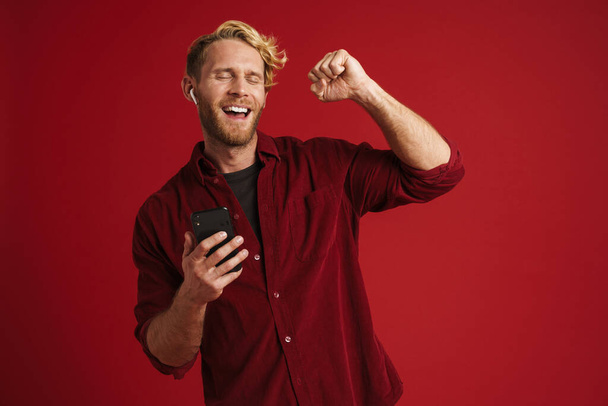 Blonde happy man listening music with earphones and dancing isolated over red wall - Foto, immagini