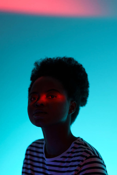 Abstraktní fotografie mladé africké ženy s neonově červeným pruhem světla na tváři izolované na živém pestrobarevném pozadí ve studiu, černé digitální dívka pózující v barevných jasných světlech, izolované na modré - Fotografie, Obrázek