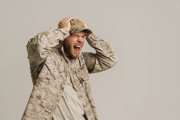 Furious military man wearing uniform holding head and screaming isolated over white wall - Zdjęcie, obraz