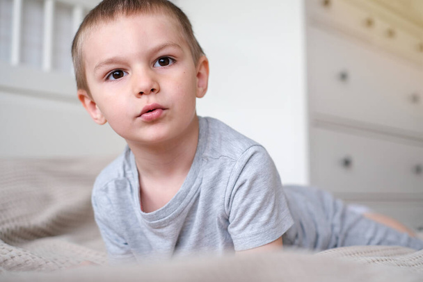 Primo piano di un piccolo ragazzo caucasico felice con una t-shirt grigia sdraiato sul letto e sorride. - Foto, immagini