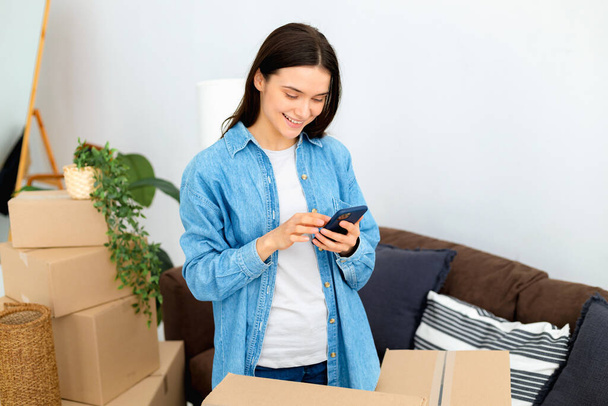 Happy young woman using mobile phone to search and order transportation service and movers to move to a new home. Moving to a new house, rental housing concept - Fotografie, Obrázek