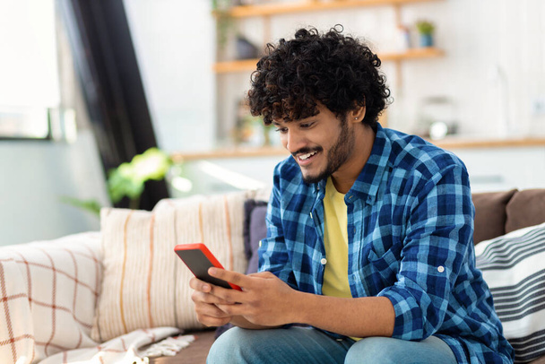 Young joyful Asian man using mobile phone and smiling sitting at home on the couch. Male holds smartphone in his hands looking at the screen, reads a message, plays games - Foto, Bild