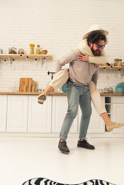 White woman in a hat piggybacking on her laughing boyfriends back having fun in the kitchen. Caucasian couple. Vertical indoor shot. High quality photo - Foto, immagini