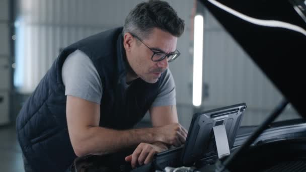 In a car service a mechanic in glasses inspecting and checking the car using a tablet computer that, standing on the opened hood of the car - Кадры, видео
