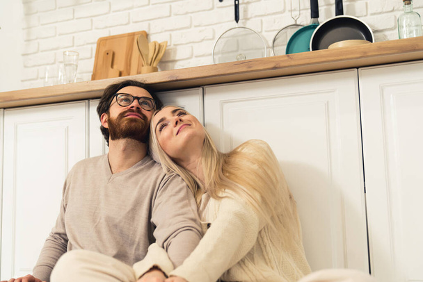 Ein junges kaukasisches Paar, das am Küchentisch auf dem Boden sitzt und Hände hält, eine blonde Frau, die sich auf einen dunkelhaarigen Mann mit Bart stützt. Indoor-Schuss. Hochwertiges Foto - Foto, Bild