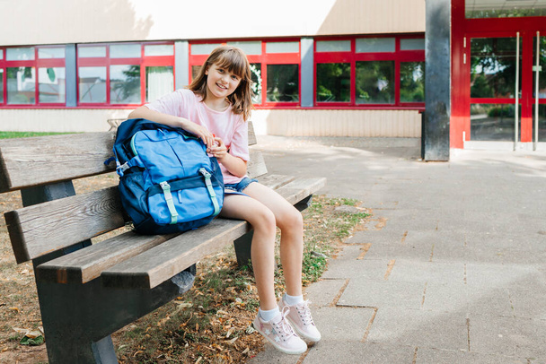 Okula dönelim. Liseli bir kız okul çantasının yanındaki bankta oturuyor, gülümsüyor ve kameraya bakıyor. - Fotoğraf, Görsel