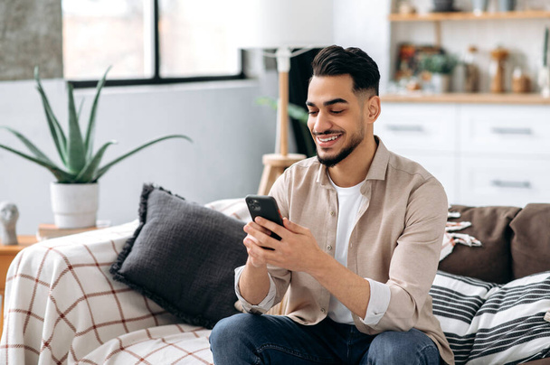 Позитивний привабливий дружній індійський або арабський хлопець, стильно одягнений, сидить на дивані в інтер'єрі вітальні, використовує свій смартфон, спілкується в соціальних мережах з друзями, переглядаючи онлайн-магазини
 - Фото, зображення