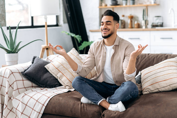 Happy positive calm relaxed indian or arabian man in casual clothes, sitting alone at home in living room on the couch and meditating in the lotus position, take care of his mental health, smiling - Foto, Imagem