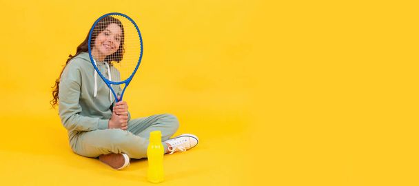 onnellinen lapsi istuu urheiluvaatteissa squash-mailalla ja vesipullolla keltaisella taustalla. Horisontaalinen juliste eristetty lapsen kasvot, banneri otsikko, kopioi tilaa - Valokuva, kuva