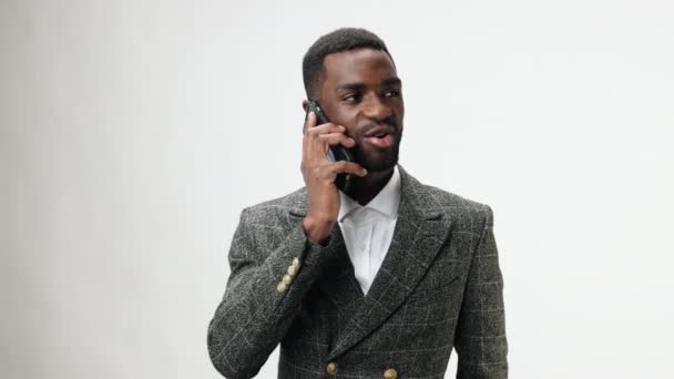 Afrikai-amerikai férfi beszél telefonon a kezében és nevet, barátságos beszélgetés szürke dzsekiben és fehér ing szürke stúdió háttérben. Kiváló minőségű 4k felvételek - Felvétel, videó