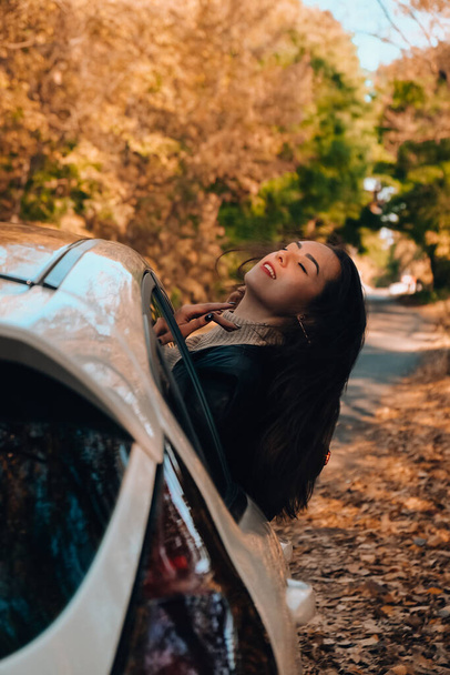 Brunette European looking cute girl sitting in a car enjoying sunny autumn weekend. Travel and road trip. Woman looking at the view from a car. Happy woman enjoying road trip. Fall Autumn forest in - Fotoğraf, Görsel