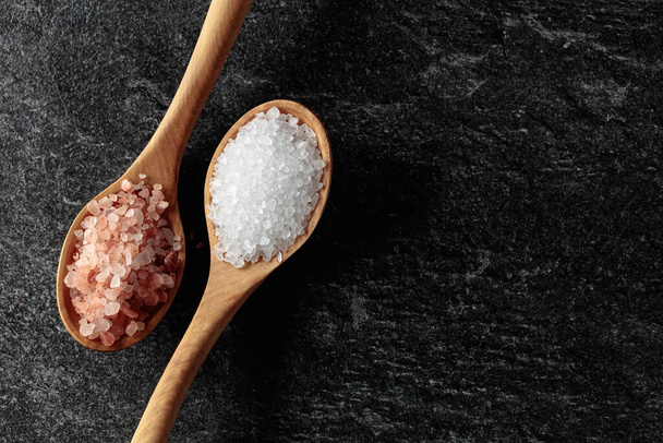 Pink and sea salt in wooden spoons on a black stone background. Copy space. - Φωτογραφία, εικόνα