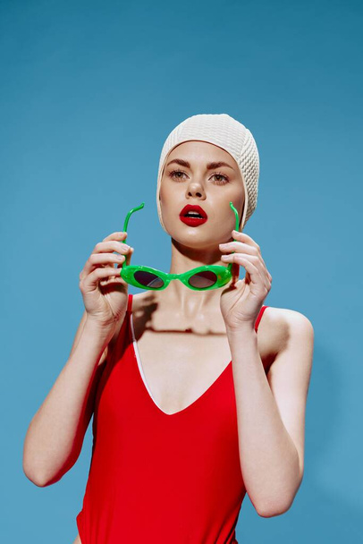 A shocked, surprised young female swimmer in a red swimsuit puts on glasses in the studio on a blue background. Art fashion. High quality photo - Foto, imagen