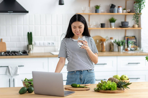 Asian young woman vlogger live broadcast cooking healthy food standing at home in the kitchen looking at laptop screen, smiling friendly - Foto, Bild