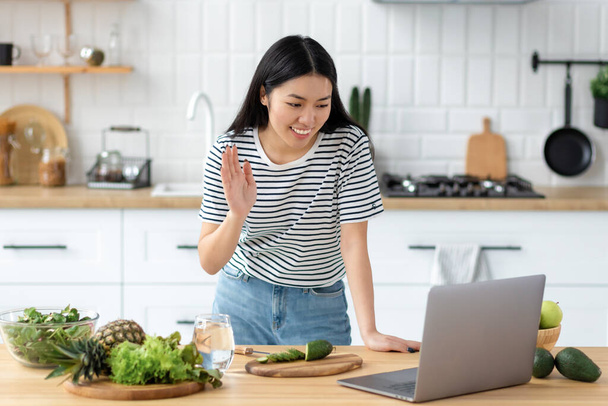 Asian young woman vlogger live broadcast cooking healthy food standing at home in the kitchen waving at the laptop, smiling friendly - Foto, afbeelding