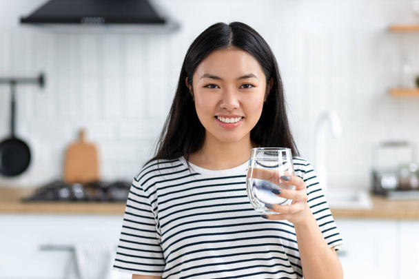 Healthy lifestyle concept. Beautiful young Asian woman holding a glass of clean fresh water standing at home in the kitchen, looks at the camera and smiles friendly - Foto, Imagem