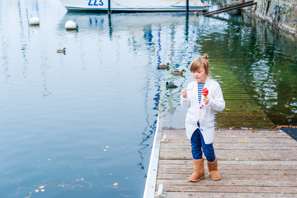 Bonita niña vestida de punto blanco cárdigan y botas de color marrón cálido, comiendo helado al lado del lago
 - Foto, Imagen