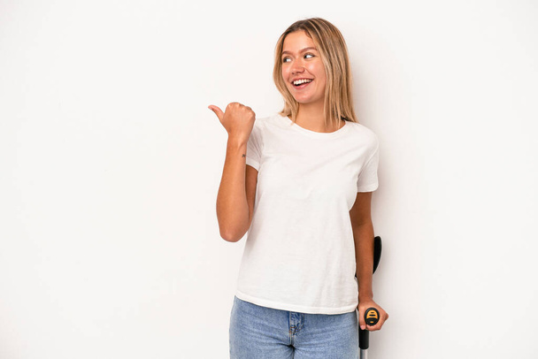 Young caucasian woman holding crutch isolated on white background points with thumb finger away, laughing and carefree. - Foto, imagen