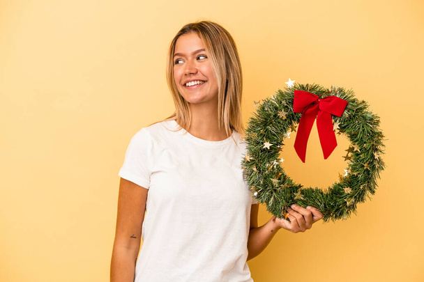 Fiatal fehér nő kezében egy karácsonyi koszorú elszigetelt sárga háttér néz félre mosolygós, vidám és kellemes. - Fotó, kép