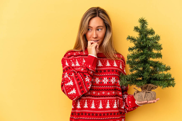 Νεαρή λευκή γυναίκα που κρατάει ένα μικρό χριστουγεννιάτικο δέντρο απομονωμένο σε κίτρινο φόντο χαλαρό σκεπτόμενη κάτι κοιτάζοντας ένα χώρο αντιγραφής. - Φωτογραφία, εικόνα