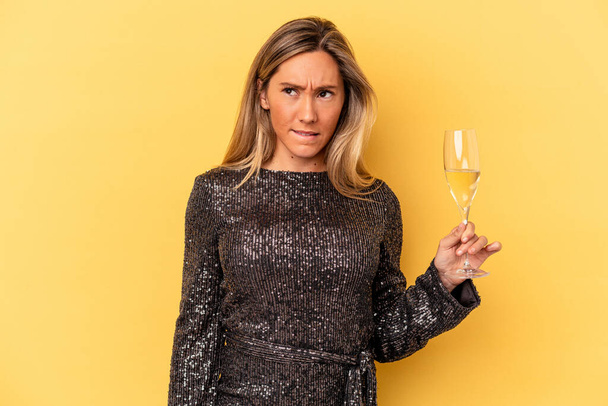 Young caucasian woman celebrating new year holding cava cup isolated on yellow background confused, feels doubtful and unsure. - Foto, Imagen