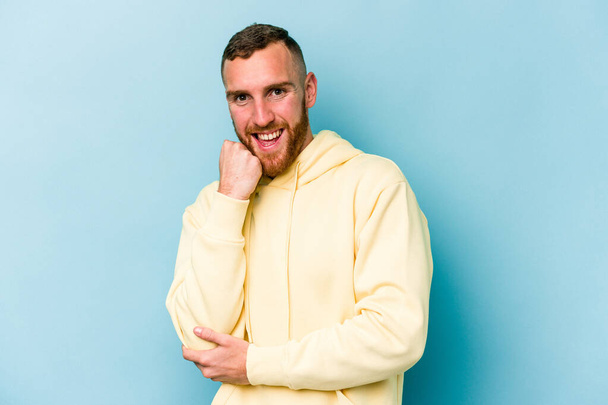 Young caucasian man isolated on blue background smiling happy and confident, touching chin with hand. - Photo, Image