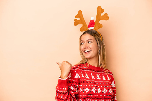 Young caucasian woman wearing a christmas reindeer hat isolated on beige background points with thumb finger away, laughing and carefree. - Foto, Imagem