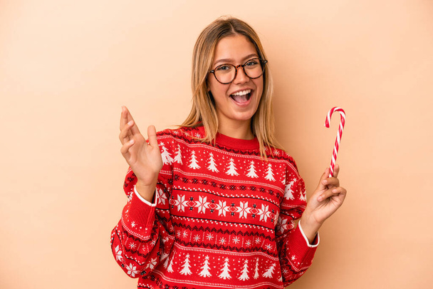 Молода біла жінка тримає різдвяну паличку ізольовано на бежевому тлі отримує приємний сюрприз, збуджує і піднімає руки
. - Фото, зображення