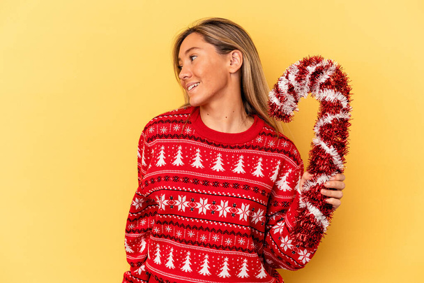 Joven mujer caucásica sosteniendo gran palo de Navidad aislado sobre fondo amarillo se ve a un lado sonriente, alegre y agradable. - Foto, imagen