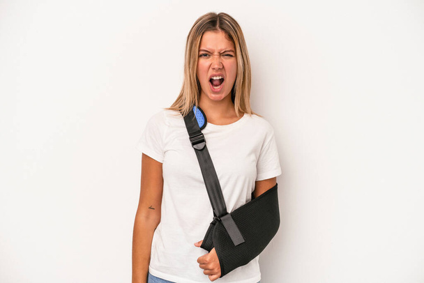 Young caucasian woman with broken hand isolated on white background screaming very angry and aggressive. - Zdjęcie, obraz