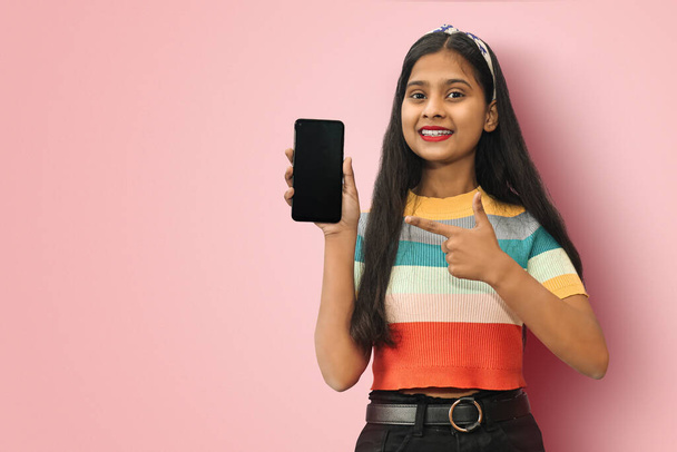 Smiling Indian Asian girl posing isolated is holding blank screen mobile phone and pointing finger to a copy space. - Foto, afbeelding