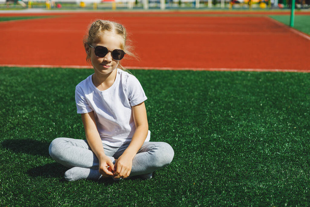 A little girl of 7-8 years old in a white T-shirt and sunglasses is sitting on the sports field. The girl is sitting on the green grass on a sunny day - 写真・画像